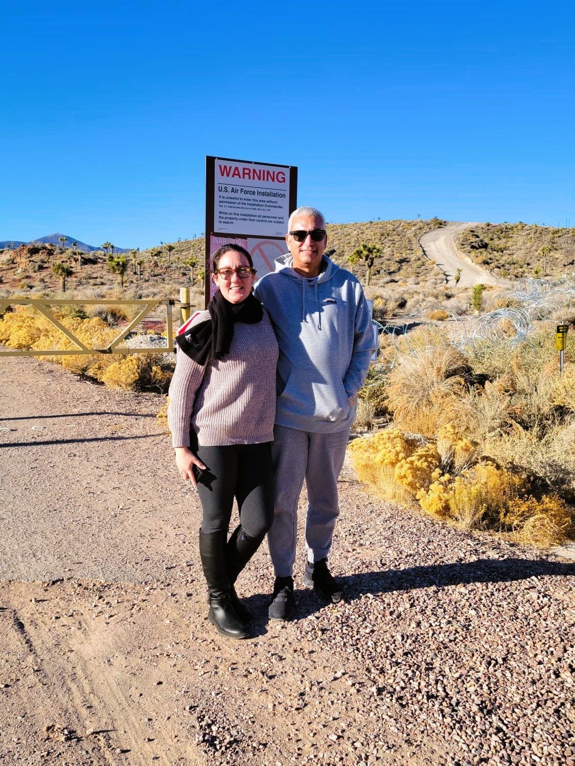 Tourists in a rugged, desert landscape with sparse vegetation, gazing towards the distant, heavily fenced perimeter of Area 51 under a vast, blue sky, Tourists in a rugged, desert landscape with sparse vegetation, gazing towards the distant, heavily fenced perimeter of Area 51 under a vast, blue sky,